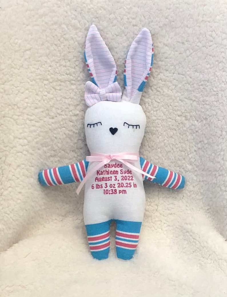 May include: White bunny plush toy with blue and pink striped legs and ears. The bunny has embroidered eyes and a pink bow. The bunny has embroidered text that reads "Saydee Kathleen Syde August 3, 2022 6 lbs 3 oz 20.25 in 10:38 pm".
