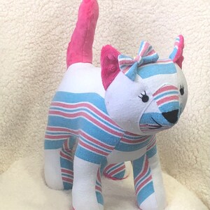 K&ouml;nnte beinhalten: Ein rosa-blaues gestreiftes Pl&uuml;schkatzenspielzeug mit einer rosa Schleife. Die Katze hat ein wei&szlig;es Gesicht mit schwarzen Augen und einer schwarzen Nase.