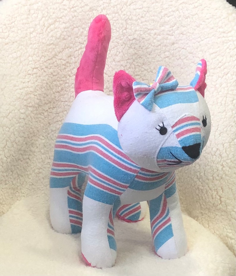 K&ouml;nnte beinhalten: Ein rosa-blaues gestreiftes Pl&uuml;schkatzenspielzeug mit einer rosa Schleife. Die Katze hat ein wei&szlig;es Gesicht mit schwarzen Augen und einer schwarzen Nase.