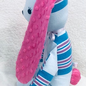 May include: A plush bunny rabbit with light blue body and pink accents. The long ears are pink with a textured surface. The bunny's body is made from fabric with blue and purple stripes. A small pink pom-pom tail completes the design.