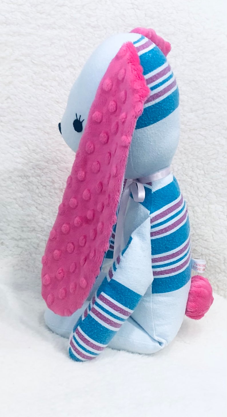 May include: A plush bunny rabbit with light blue body and pink accents. The long ears are pink with a textured surface. The bunny's body is made from fabric with blue and purple stripes. A small pink pom-pom tail completes the design.