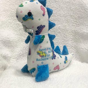 May include: A plush dinosaur toy in white fabric with blue accents. The dinosaur has blue spikes, feet, and a tail. It is decorated with colorful shapes and has embroidered text: David York, April 15, 2024, 9.8 lbs, Rochester, NY.