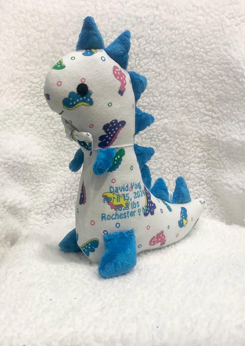 May include: A plush dinosaur toy in white fabric with blue accents. The dinosaur has blue spikes, feet, and a tail. It is decorated with colorful shapes and has embroidered text: David York, April 15, 2024, 9.8 lbs, Rochester, NY.