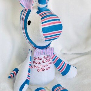 May include: A white unicorn stuffed animal with blue and pink striped fabric. The unicorn has a bow tie and is embroidered with the text "Th&eacute;o Smi Aug. 4, 2021 6 lbs. 11 oz. 19 in 8:05 am".