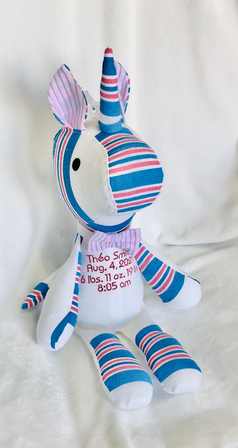 May include: A white unicorn stuffed animal with blue and pink striped fabric. The unicorn has a bow tie and is embroidered with the text "Th&eacute;o Smi Aug. 4, 2021 6 lbs. 11 oz. 19 in 8:05 am".