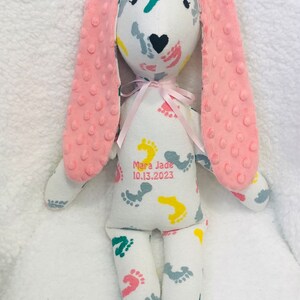 May include: A white bunny plush toy with pink ears and a pink bow. The bunny's body is covered in colorful footprints. The text "Mara Jade 10.13.2023" is printed on the bunny's body.