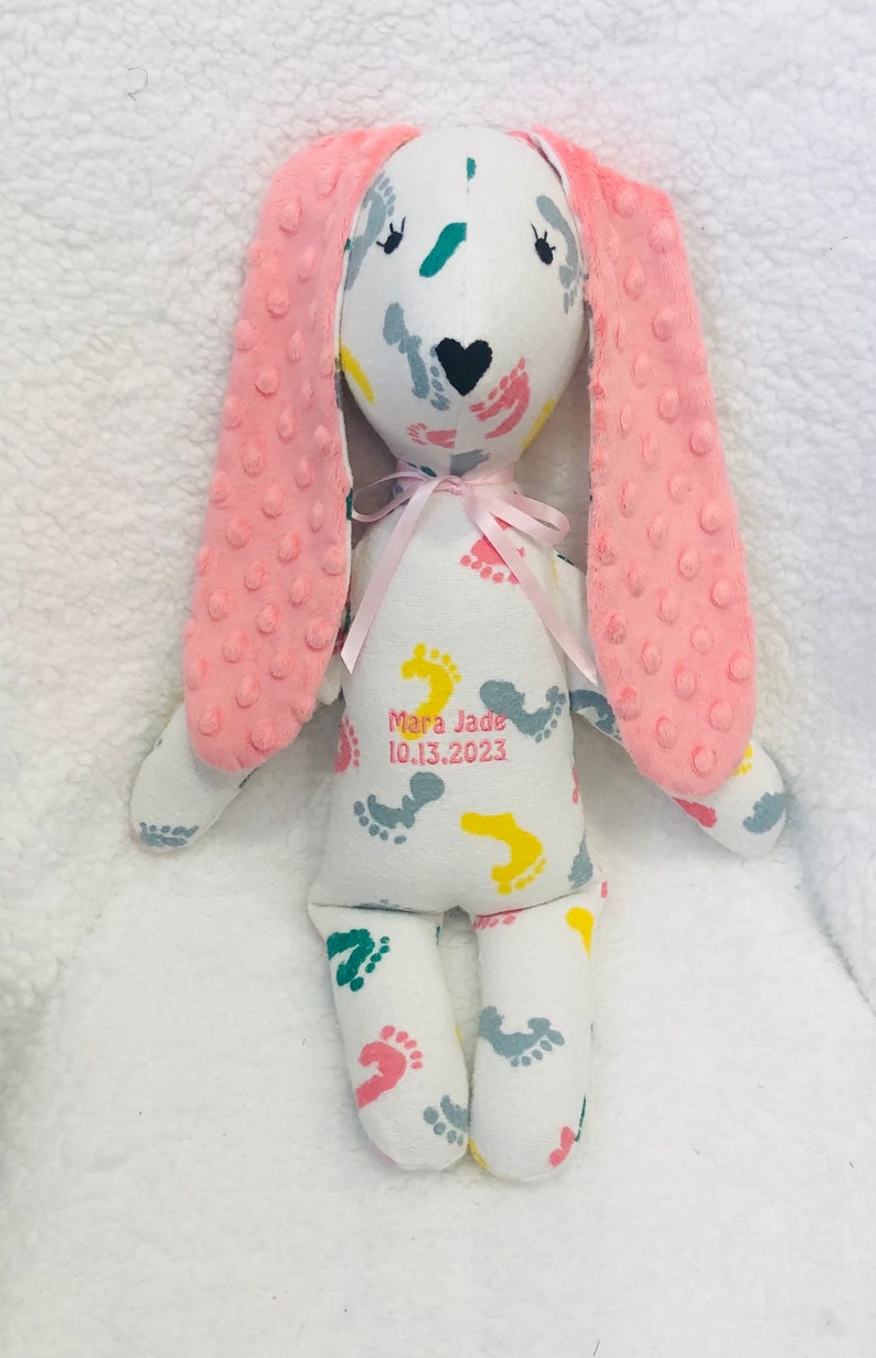 May include: A white bunny plush toy with pink ears and a pink bow. The bunny's body is covered in colorful footprints. The text "Mara Jade 10.13.2023" is printed on the bunny's body.