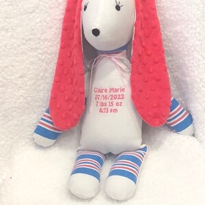 May include: A plush bunny doll with a white body, red textured ears, and blue and white striped accents on the arms and legs. The doll has embroidered facial features and a pink ribbon. Text on the body reads "Claire Marie 07/16/2022 7 lbs 15 oz 6:13 pm."