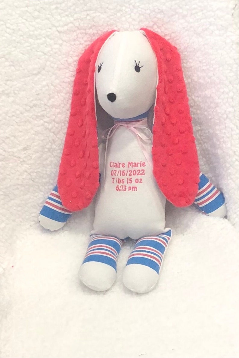 May include: A plush bunny doll with a white body, red textured ears, and blue and white striped accents on the arms and legs. The doll has embroidered facial features and a pink ribbon. Text on the body reads "Claire Marie 07/16/2022 7 lbs 15 oz 6:13 pm."