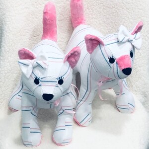 K&ouml;nnte beinhalten: Zwei Pl&uuml;schkatzen mit rosa Ohren und Schw&auml;nzen. Beide Katzen sind wei&szlig; mit rosa und blauen Streifen und haben eine rosa Schleife auf dem Kopf.