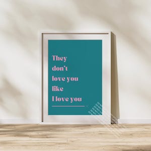 May include: A framed art print with the text "They don't love you like I love you" in pink against a teal background. The print is in a white frame and leans against a wall.