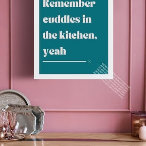 May include: A framed teal print with the text "Remember cuddles in the kitchen, yeah" in white. The print hangs on a pink wall above a wooden countertop and pink cabinets. Silver serving trays and glassware are on the counter.