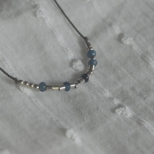 Puede incluir: Un collar delicado con una cadena de plata y cuentas azules. El collar está sobre un fondo de tela blanca.