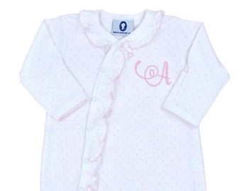 May include: A white long-sleeved footed sleeper with a delicate pink polka dot pattern. The garment features a ruffled trim along the asymmetrical snap closure and a pink embroidered letter A on the chest. A blue brand label reads Anne Everett.