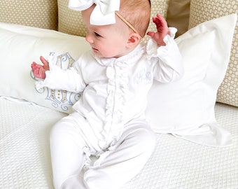 May include: A baby wearing a white ruffled onesie with a blue embroidered monogram. The baby is wearing a white bow headband and is sitting on a white quilted bedspread.