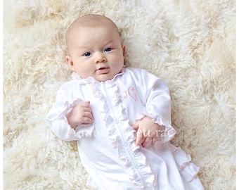 May include: A white baby sleeper with pink ruffles and a monogrammed "JG" on the chest. The baby is lying on a white fluffy rug.