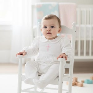 May include: A white long-sleeved infant romper featuring a Peter Pan collar, a pink embroidered cross on the chest, and a delicate lace waistband, worn by a baby sitting in a white wooden rocking chair with a high back and curved armrests.