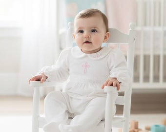 May include: A white long-sleeved infant romper featuring a Peter Pan collar, a pink embroidered cross on the chest, and a delicate lace waistband, worn by a baby sitting in a white wooden rocking chair with a high back and curved armrests.