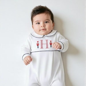 May include: A white long-sleeved footed sleeper with a Peter Pan collar featuring dark navy piping. The chest displays a red embroidered letter H flanked by two small red and navy nutcracker figures. The garment has a pleated front bodice.