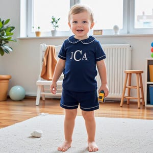 May include: A navy blue short-sleeve shirt and matching shorts set featuring white piping trim along the collar, sleeves, and hem. The shirt displays the white embroidered monogram letters JCA centered on the chest in a classic serif font.
