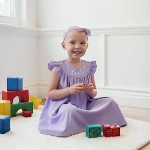 May include: A lavender dress with ruffle sleeves and a matching headband. The dress has the name "Leslie" embroidered on the front. Colorful wooden blocks and toys are scattered around the child.