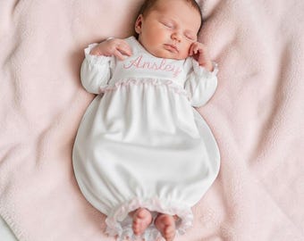 May include: A white infant gown with long sleeves, ruffled cuffs, and a ruffled waistline. The chest features the name Ansley embroidered in pink script. The garment is made of a smooth fabric and is designed with an open bottom for easy access.