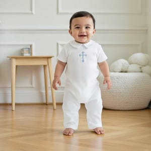 May include: A white short-sleeved baby romper featuring a Peter Pan collar and a light blue embroidered cross centered on the chest. The garment has a simple, clean design with full-length legs and a classic round neckline for a comfortable fit.