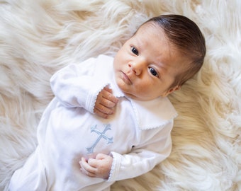 May include: A white long-sleeved infant gown featuring a pointed collar with scalloped edges and a light blue embroidered cross on the chest. The garment is made of a smooth fabric and includes long sleeves with matching scalloped cuffs at the wrists.