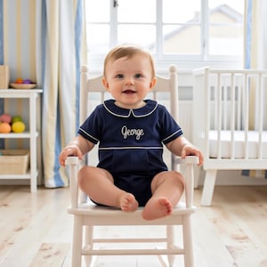 May include: A navy blue short-sleeved romper with a white Peter Pan collar and white piping trim. The romper features the name George embroidered in white script on the chest. The garment has a pleated front detail and short leg openings.