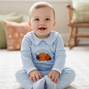 May include: A light blue and white gingham footed sleeper with a Peter Pan collar and long sleeves. The chest features an embroidered applique of a football, a large orange pumpkin, a smaller orange pumpkin, and a brown acorn with green leaves.