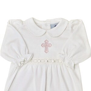 May include: A white long-sleeved gown featuring a rounded Peter Pan collar with scalloped edges, gathered sleeve cuffs, and a horizontal band of white eyelet lace trim at the waist. A pink embroidered cross is centered on the chest area.