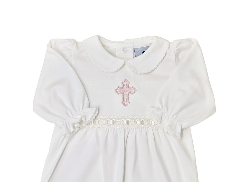 May include: A white long-sleeved gown featuring a rounded Peter Pan collar with scalloped edges, gathered sleeve cuffs, and a horizontal band of white eyelet lace trim at the waist. A pink embroidered cross is centered on the chest area.