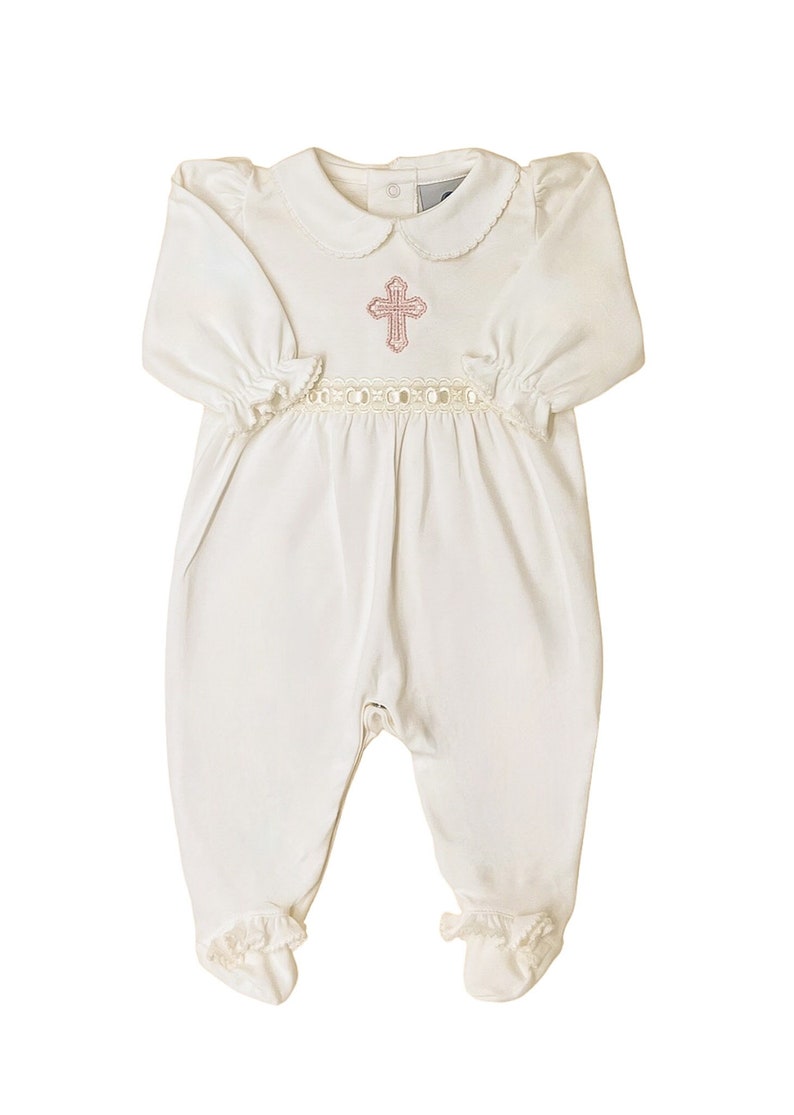 May include: A white long-sleeved footed romper featuring a rounded collar with scalloped edges, a pink embroidered cross on the chest, and a decorative cream-colored lace trim at the waist. The sleeves and feet have gathered elasticized cuffs.