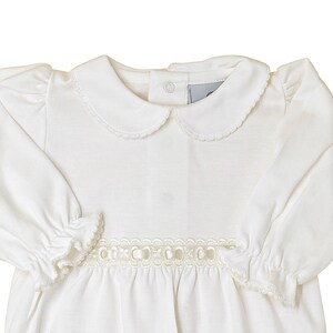 May include: A long-sleeved white dress featuring a rounded Peter Pan collar with delicate scalloped edges, gathered puff shoulders, and elasticized ruffled cuffs. A decorative cream-colored ribbon trim with circular cutouts accents the waist.