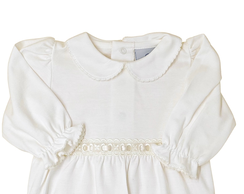 May include: A long-sleeved white dress featuring a rounded Peter Pan collar with delicate scalloped edges, gathered puff shoulders, and elasticized ruffled cuffs. A decorative cream-colored ribbon trim with circular cutouts accents the waist.
