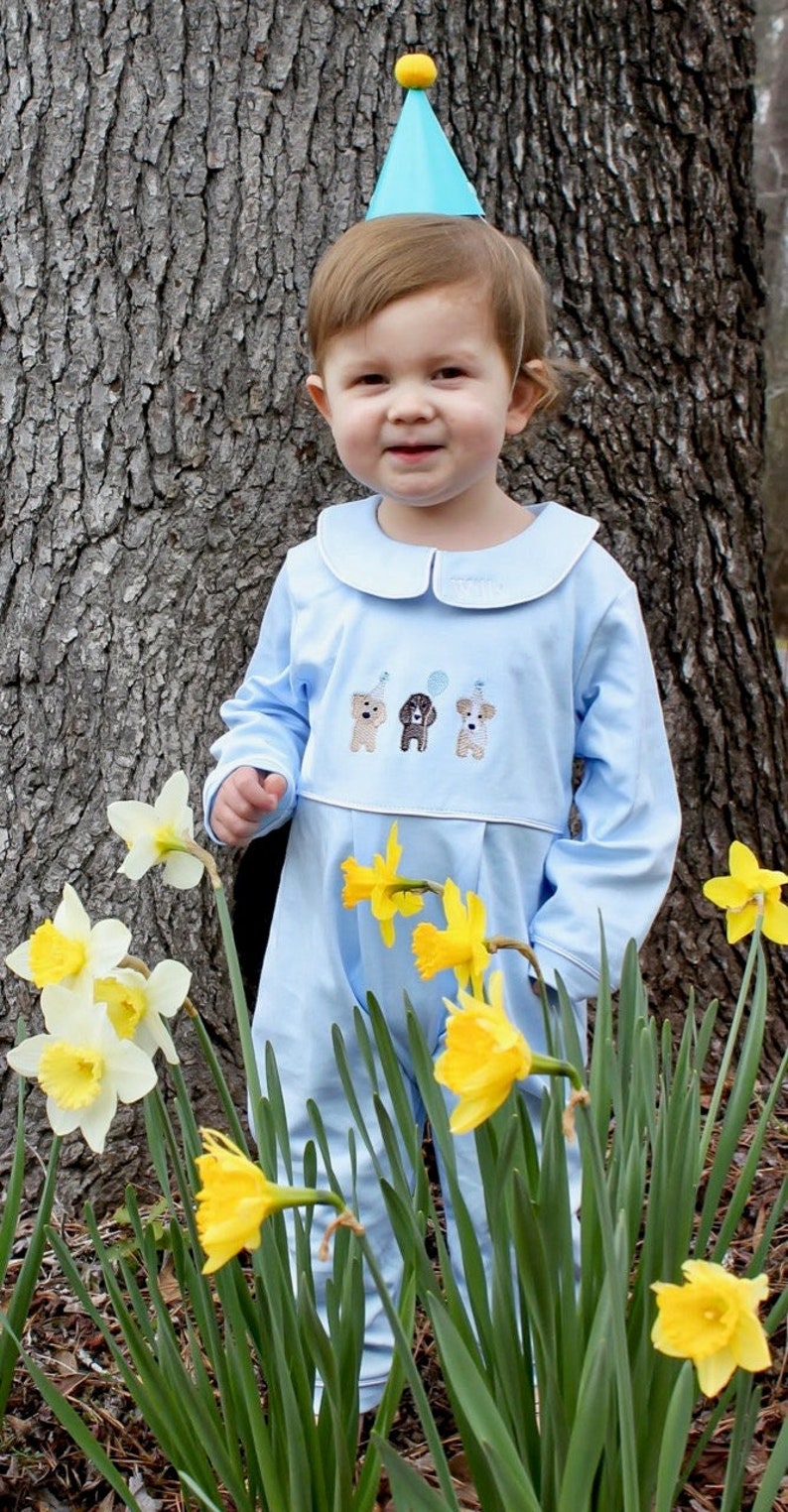 May include: A young child wearing a blue outfit with a white collar and three embroidered dogs on the front. The child is wearing a blue and yellow party hat and is standing in front of a tree with yellow daffodils.