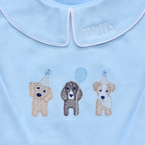 May include: A light blue long-sleeved shirt with a white collar and the name "Wills" embroidered on the collar. Three embroidered dogs wearing party hats are on the shirt, one holding a balloon.