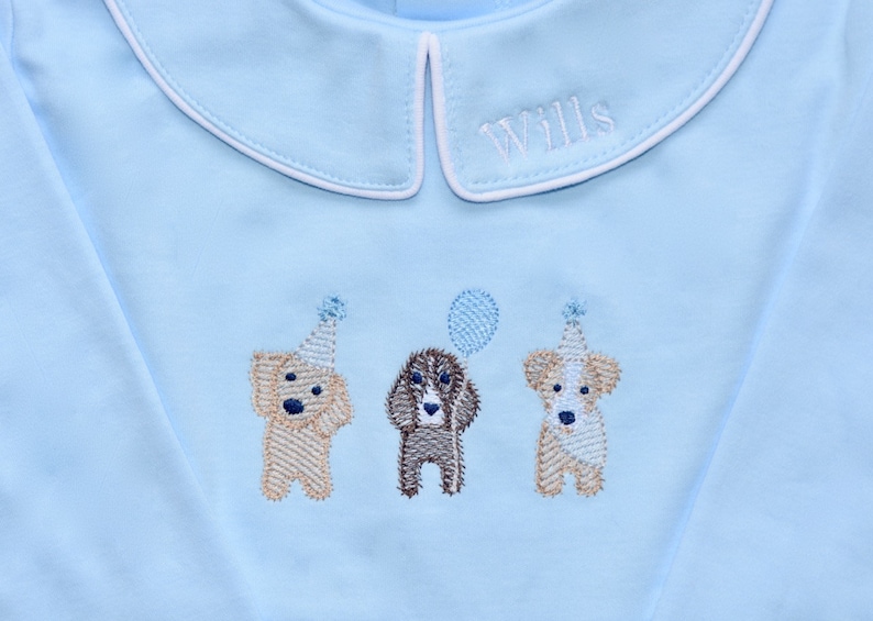 May include: A light blue long-sleeved shirt with a white collar and the name "Wills" embroidered on the collar. Three embroidered dogs wearing party hats are on the shirt, one holding a balloon.