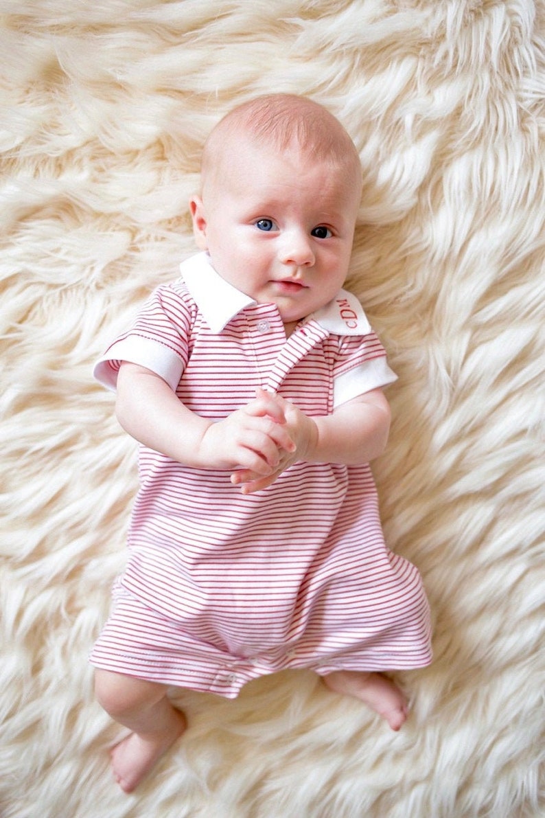 May include: A baby wearing a pink and white striped onesie with a white collar and red embroidered initials "CDD". The baby is lying on a white fluffy rug.