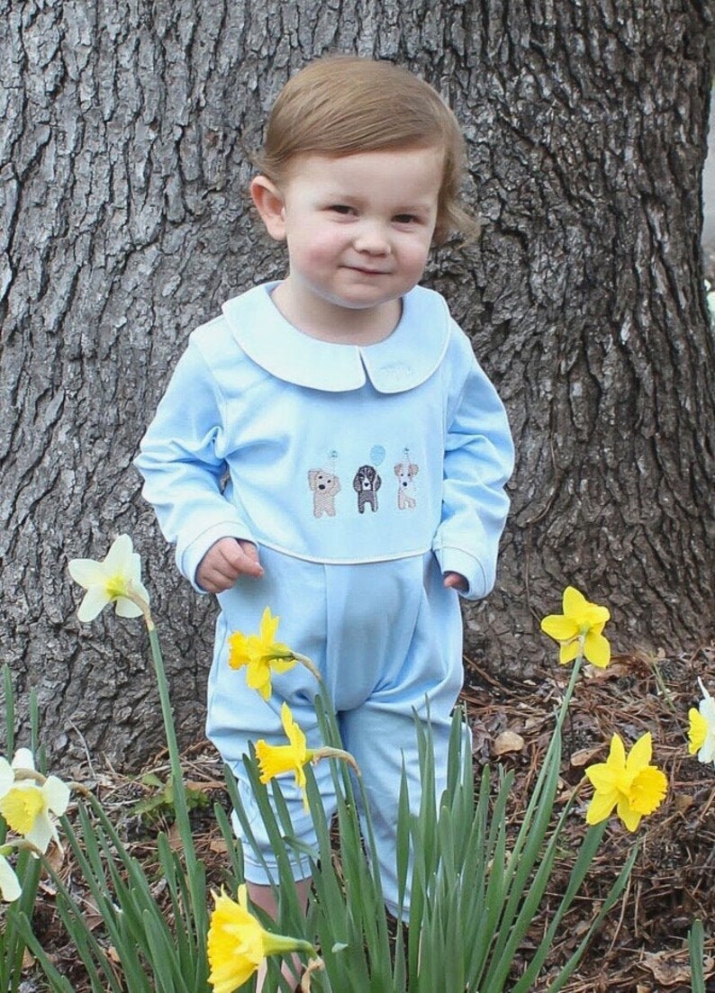 May include: A young child wearing a light blue long-sleeved jumpsuit with a white collar and embroidered dogs. The jumpsuit has a white band around the waist and a white band at the bottom of the legs. The child is standing in a grassy area with yellow daffodils.
