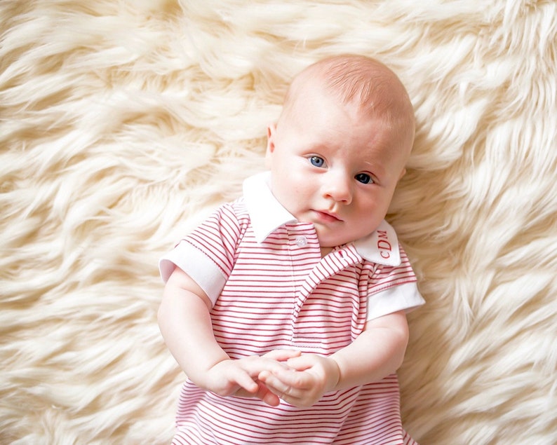 May include: A baby wearing a red and white striped onesie with a white collar and the letters "CDM" embroidered on the collar.