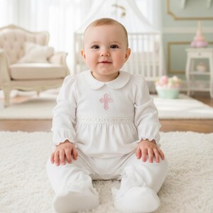 May include: A white long-sleeved infant footed sleeper featuring a rounded collar with scalloped edges, a delicate lace waistband, and a small pink embroidered cross centered on the chest. The garment has gathered sleeves and integrated foot covers.