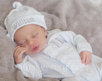 May include: A white newborn sleeper and matching knotted hat with a light blue polka dot pattern. The hat features the embroidered name William in grey thread, and the sleeper displays the embroidered initials WR in grey on the chest area.