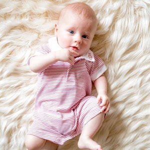 May include: A baby wearing a pink and white striped onesie with a white collar and the letters "CDM" embroidered on the collar. The baby is lying on a white fluffy rug.