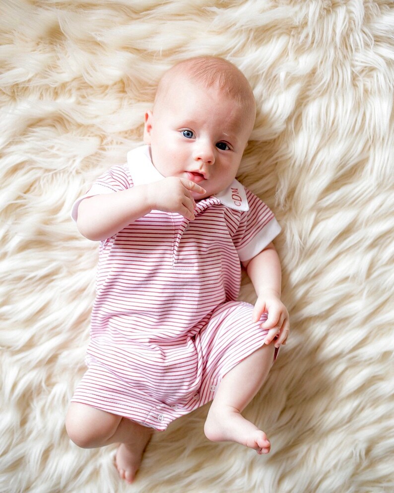 May include: A baby wearing a pink and white striped onesie with a white collar and the letters "CDM" embroidered on the collar. The baby is lying on a white fluffy rug.