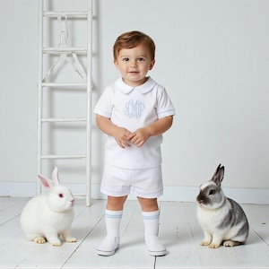 May include: A white short-sleeved shirt with a Peter Pan collar and light blue monogram embroidery, paired with matching white shorts featuring light blue trim. The outfit includes white socks with light blue stripes and white slip-on shoes.