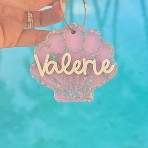 May include: A light purple seashell-shaped acrylic keychain featuring the name Valerie in white script lettering. The edges of the shell are decorated with multi-colored glitter, and the piece is attached to a silver-toned metal wire loop.