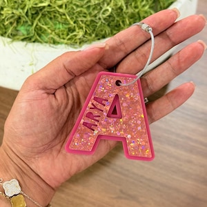 Może przedstawiać: Różowa zawieszka do bagażu w kształcie litery "A" z imieniem "ARYA" w różowych literach. Zawieszka jest wypełniona brokatem i ma srebrną pętlę drucianą do mocowania.