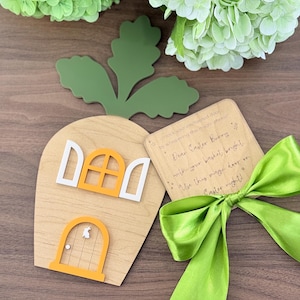 May include: Wooden carrot-shaped Easter door with an orange door and window frames. A green leaf and a card with text are next to it, tied with a green satin ribbon. The text on the card reads "Dear Easter Bunny..."