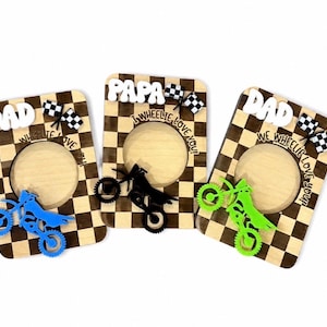 May include: Three rectangular wooden photo frames with a checkered pattern. Each frame features a cutout circle, a small checkered flag, and a motorcycle silhouette in blue, black, or green. Text reads DAD or PAPA and WE WHEELIE LOVE YOU.